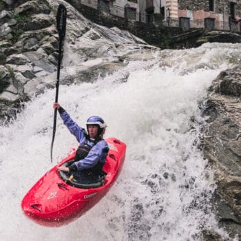 Action shot of Ava Christensen mid-air, maneuvering a red Dagger whitewater kayak over a drop in Italy.