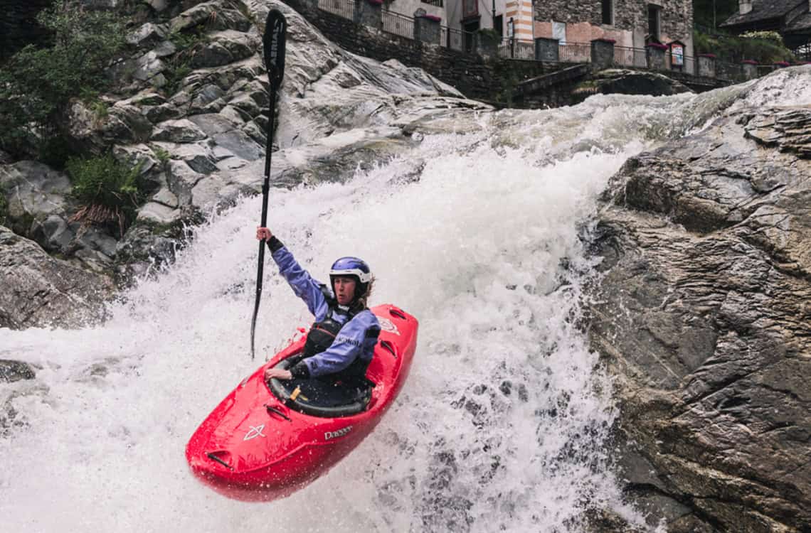 Action shot of Ava Christensen mid-air, maneuvering a red Dagger whitewater kayak over a drop in Italy.