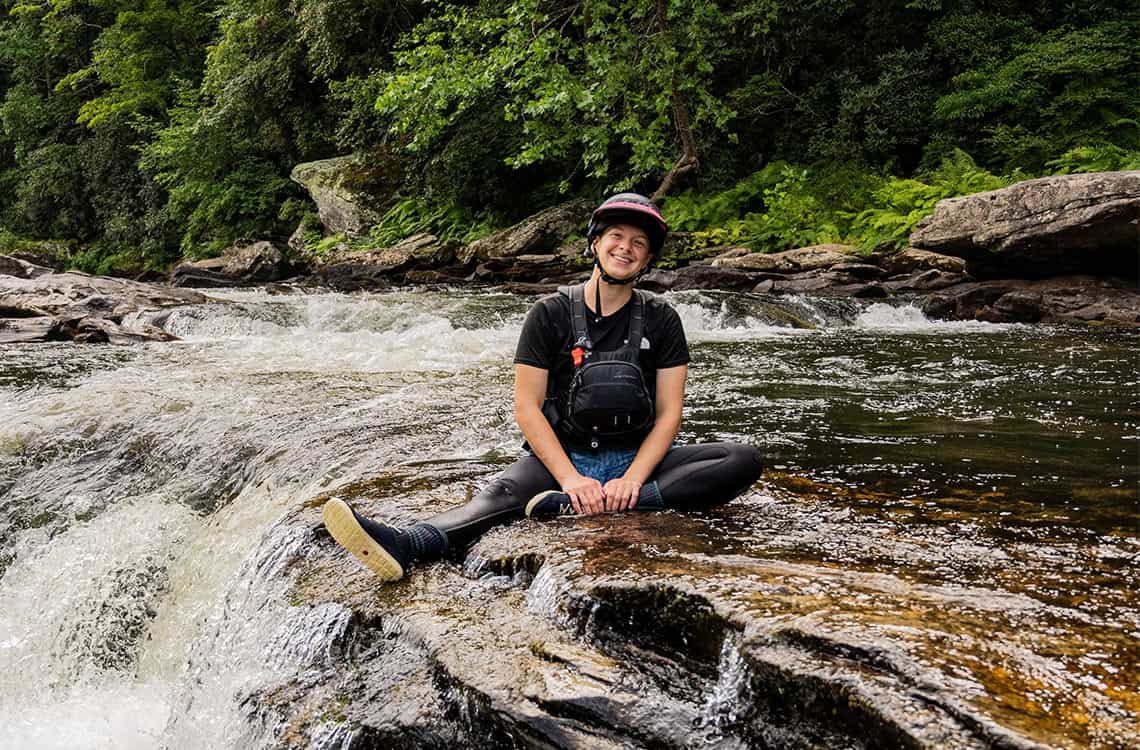 Profile photo of Danielle Sartori sitting in whitewater kayaking gear by the Chattooga River.