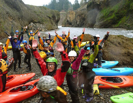 A large group of kayakers smiling with their arms up along the river