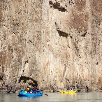 Two rafts float at the base of a towering vertical cliff wall on an Owyhee River rafting trip.