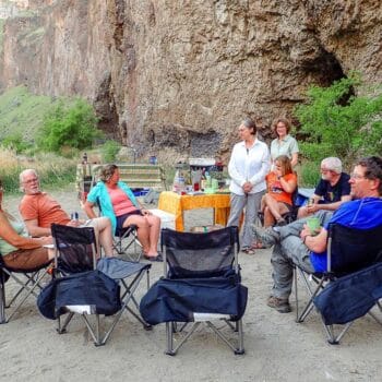 A group of rafters at camp during an Owyhee River rafting trip sit in chairs in a circle, while laughing and drinking wine.