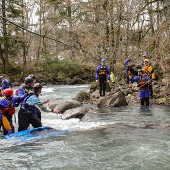 A group of students on a swiftwater river rescue course stand in the river behind a pinned kayak and wait to receive a rescue rope from shore.