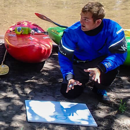 A student on the ACA kayak instructor course presents a lesson plan while kneeling next to the river.