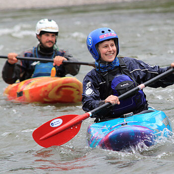 A female kayaker paddles her kayak and smiles with another kayaker in the background - Washington and Oregon river kayaking instruction and lessons.