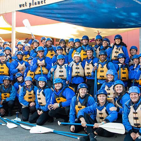 A large group of rafters in wetsuits, pfd's, and helmets poses in front of the Wet Planet Rafting and Kayaking building.