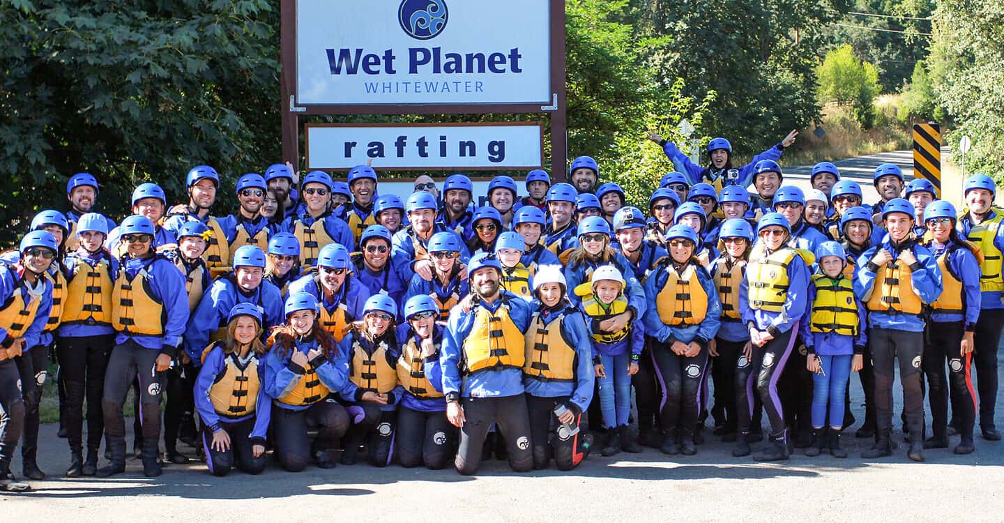 A large rafting group poses under the Wet Planet Rafting and Kayaking sign wearing their wetstuits, pfd's, and helmets.
