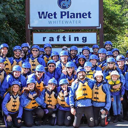 A large rafting group poses under the Wet Planet Rafting and Kayaking sign wearing their wetstuits, pfd's, and helmets.