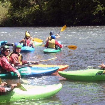 Several kayak students on a youth kayaking program sit in their kayaks on the edge of the river and listen to an instructor explain kayaking technique.