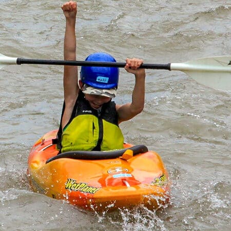 A young kayaker on the kids' kayaking camp paddles his kayak and celebrates with his fist raised in the air.