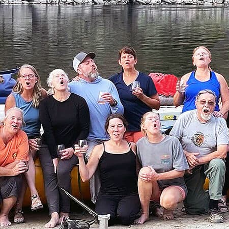 A group on a private Main Salmon river rafting trip poses in front of the rafts and makes silly faces with wine glasses in their hands.