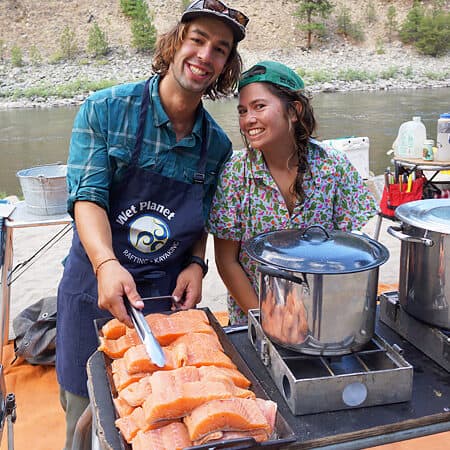 Two guides lean into each other and smile for the camera while cooking fresh salmon on a private Main Salmon river rafting trip.