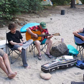 Four rafting trip guests play guitars on a beach at camp on a multi-day rafting trip.