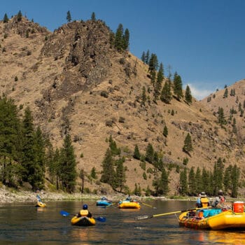A scenic shot of the Main Salmon river in Idaho with rafts and inflatable kayaks floating in calm water in front of the river canyon walls.