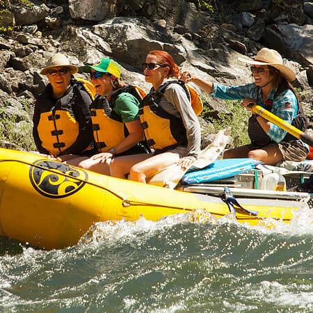 A female raft guide rows a raft through a rapid on a Mai Salmon river rafting trip while three guests smile in the front of the raft.