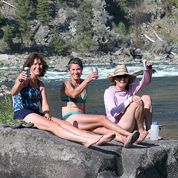 Three women relax alongside the river and raise their drinks to the camera on a Main Salmon River rafting trip in Idaho with Wet Planet whitewater