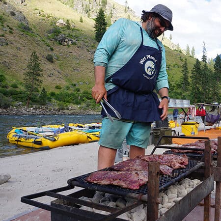 A raft guide cooks a steak meal over the grill with rafts in the background on Idaho's Main Salmon River rafting trip.
