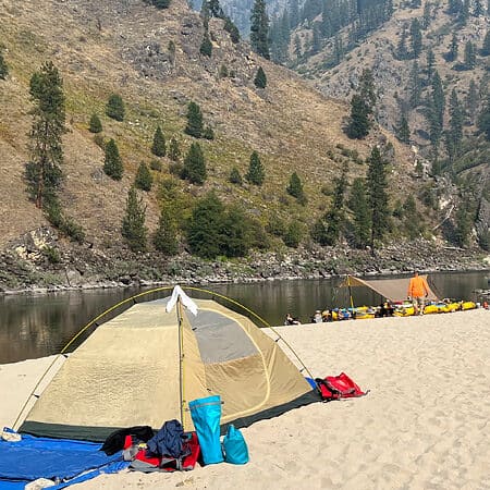 A tent is set up on a large sandy beach with rafts in the background on the Main Salmon River rafting trip in Idaho.