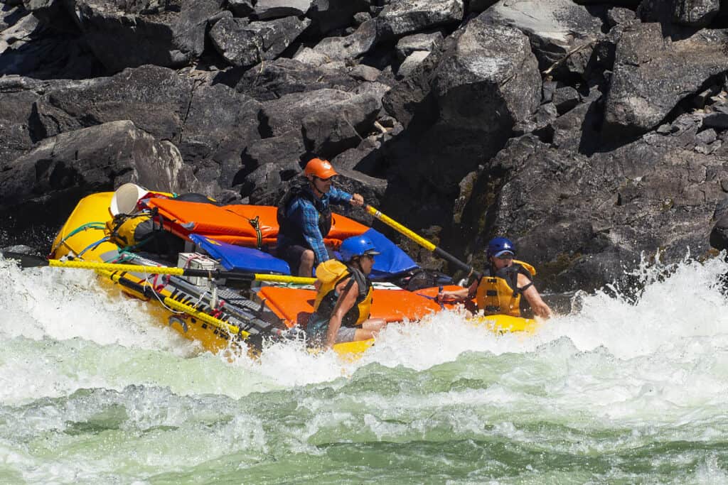 A raft guide rows a raft through a large rapid on the Main Salmon River rafting trip in Idaho while two rafters yell in excitement.