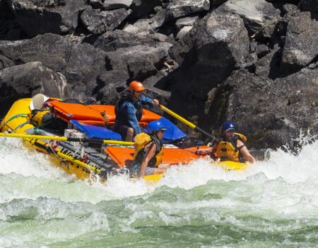 A raft guide rows a raft through a large rapid on the Main Salmon River rafting trip in Idaho while two rafters yell in excitement.