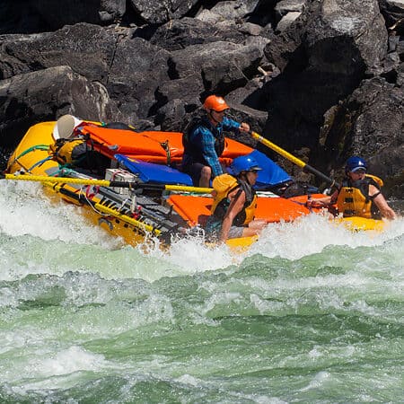 A student on an Idaho Main Salmon River rowing school rows a gear raft through a rapid with two passengers.