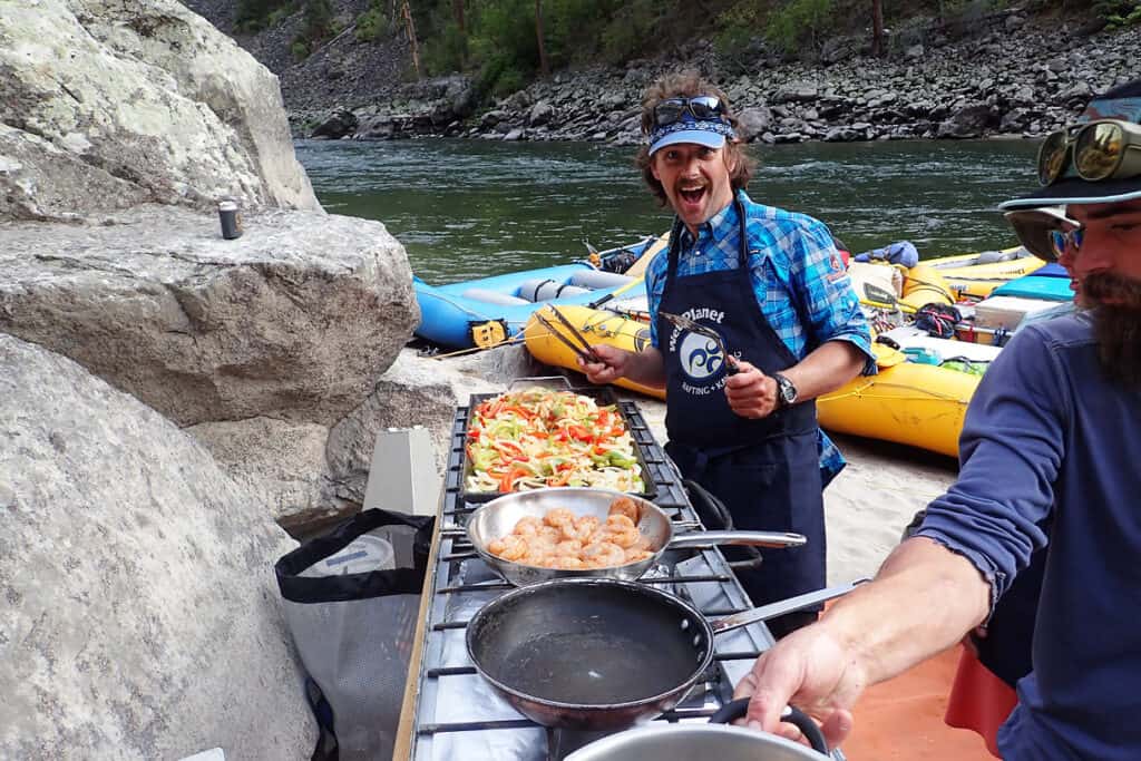 An instructor wearing a Wet Planet apron smiles at the camera while cooking breakfast in the kitchen camp.