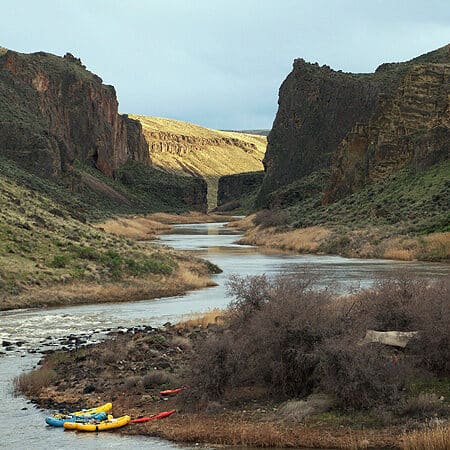 A scenic view from a rafting trip on the Owyhee River shows a group of rafts at camp with towering red canyon walls in the background.