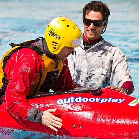 A male kayak instructor stands in the pool and watches a student prepare to do a wet exit during a kayak instruction course.