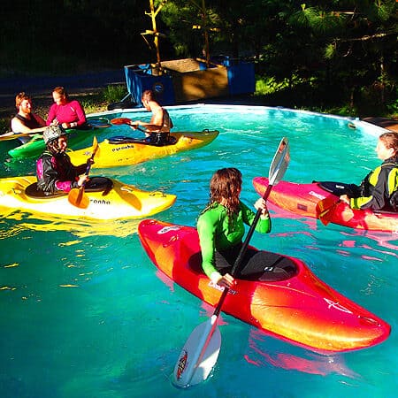 Four kayakers float in the pool and practice kayak rolls during an instruction course pool session.