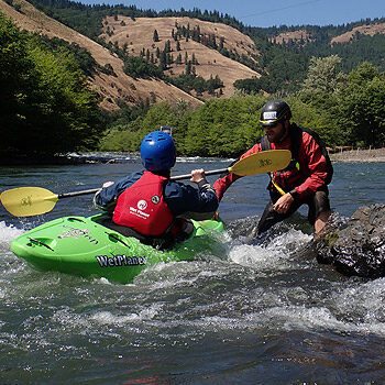 A kayak instructor stands in the water and helps a student in a kayak learn to surf on a Washington private kayak instruction course.