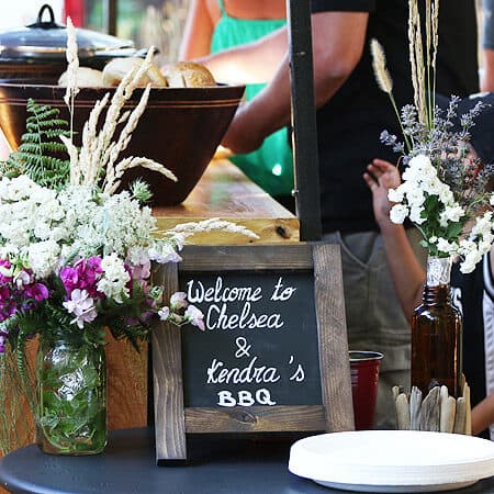 A sign welcoming guests to a BBQ is located between two vases of flowers.