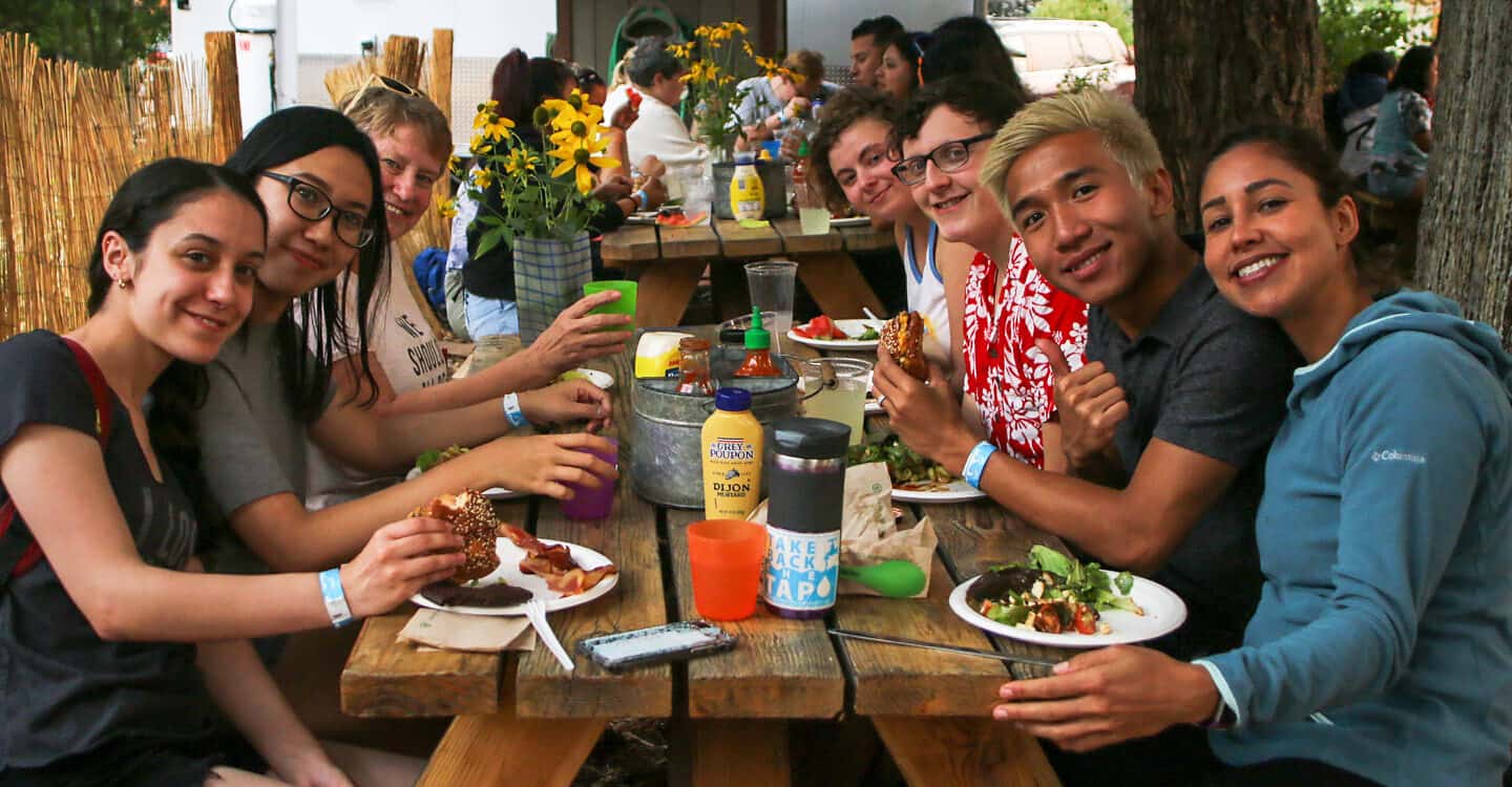 A group sits at a picnic table and smiles at the camera while eating burgers during a Wet Planet catered BBQ.