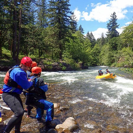 Two students on a swiftwater river rescue course belay a raft in the river with ropes.