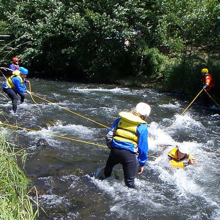 An instructor simulates a foot entrapment in a creek while students attempt to stabilize him with ropes on a swiftwater river rescue course.