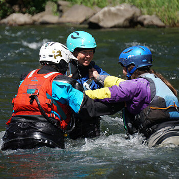 Three students on a river rescue WRT course practice shallow water wading by physically supporting each other in the current.