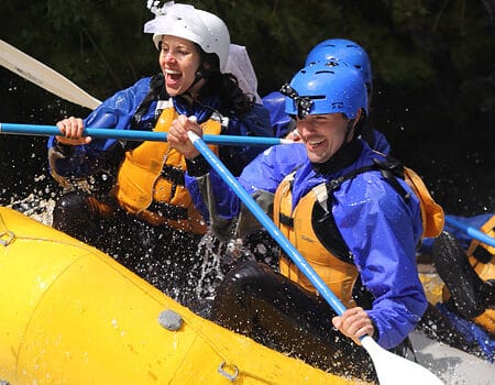 Two rafters wearing matching bride and groom helmets smile while rafting through a rapid.