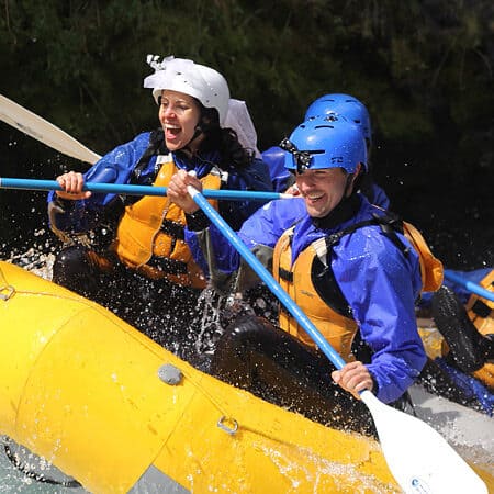 Two rafters wearing matching bride and groom helmets smile while rafting through a rapid.