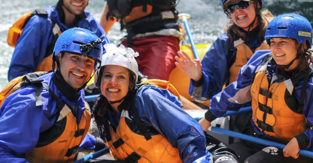 A couple on a wedding party rafting trip smiles while wearing helmets decorated with a bow tie and a bridal veil.