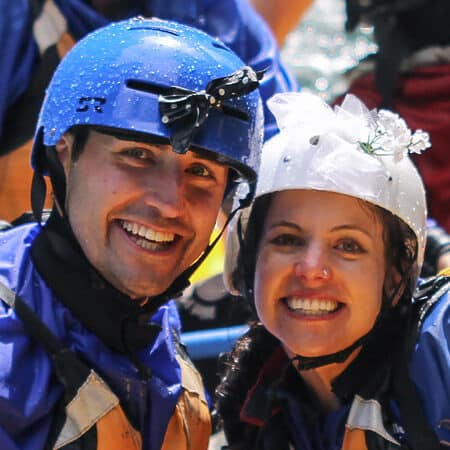 A couple on a wedding party rafting trip smiles while wearing helmets decorated with a bow tie and a bridal veil.
