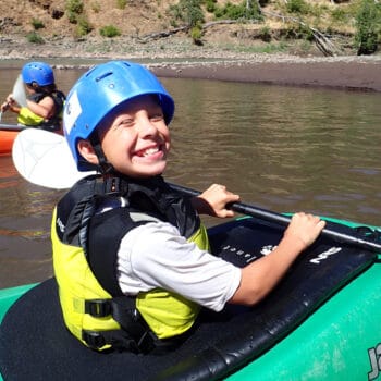 A young kayaker on a kids kayak camp sits in his kayak and shows a large smile to the camera.