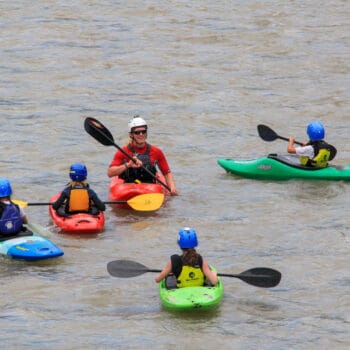 A kayak instructor watches four kids on a kayak camp paddle in flatwater.