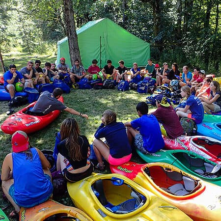 A large group of teenagers sit in a circle on top of their kayaks and watch an instructor demonstrate how to sit correctly in a river kayak.